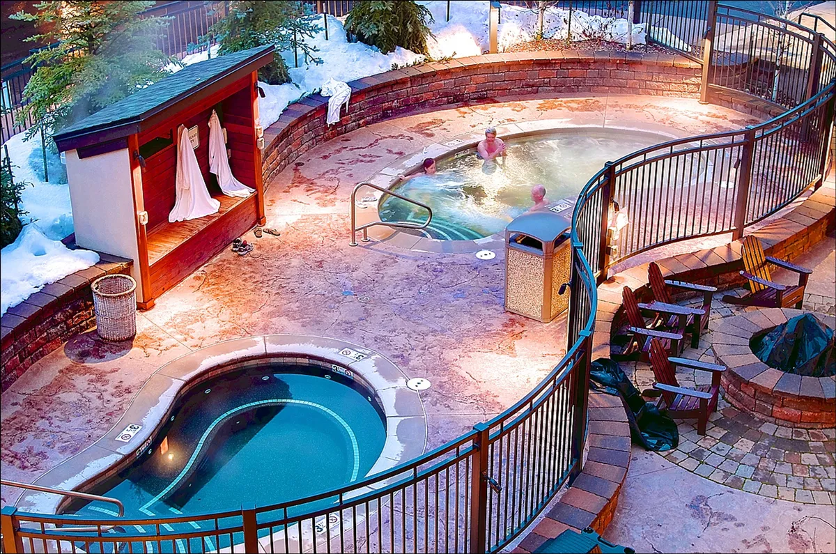 An inviting outdoor area featuring multiple hot tubs, seating, snow-covered surroundings, and decorative lighting. The scene exudes warmth and camaraderie against a classic winter backdrop.