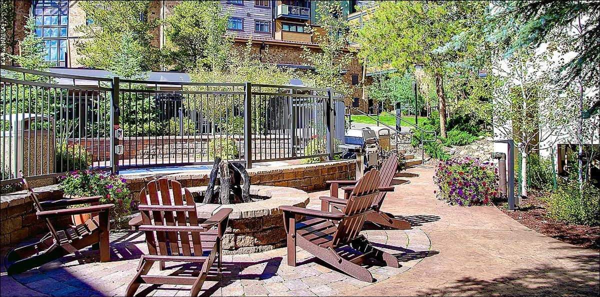 The image showcases an inviting outdoor fire pit area surrounded by Adirondack chairs set amid lush greenery, all enclosed by a rustic fence. The scene radiates warmth and relaxation, perfect for social gatherings or unwinding after a day of mountain adventure.