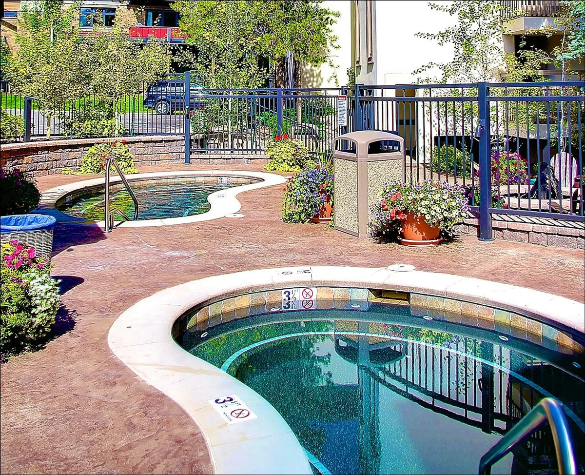 This exterior image features inviting hot tubs set amidst lush greenery and vibrant decorative flowers, all enclosed within a charming fence. The scene is tranquil and outdoorsy, highlighting the proximity of relaxation and nature just steps from the property.