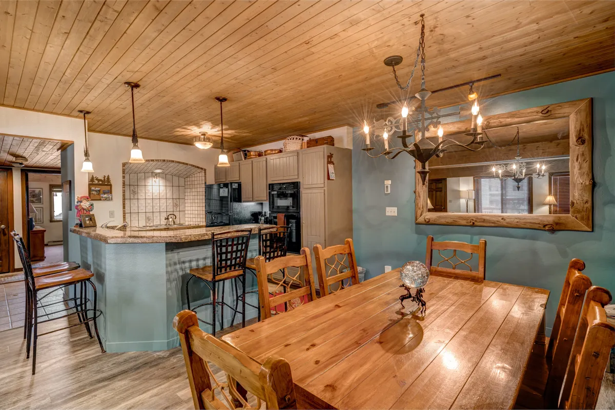 This image focuses on the dining area, showcasing a large wooden dining table with ample seating, a stylish chandelier overhead, and a bar counter visible nearby. The area is set for a cozy and inviting meal, perfect for family gatherings or celebratory dinners after a day of adventure.