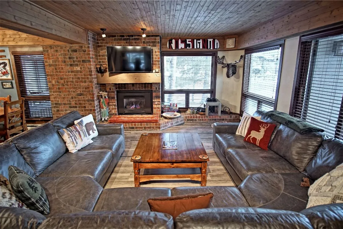 A welcoming living room centered around a leather sectional sofa and a wooden coffee table. A large fireplace anchors the space, and the decor includes tasteful wall art and a mounted TV. The big windows in the background hint at possible mountain views and easy access to the outdoors.