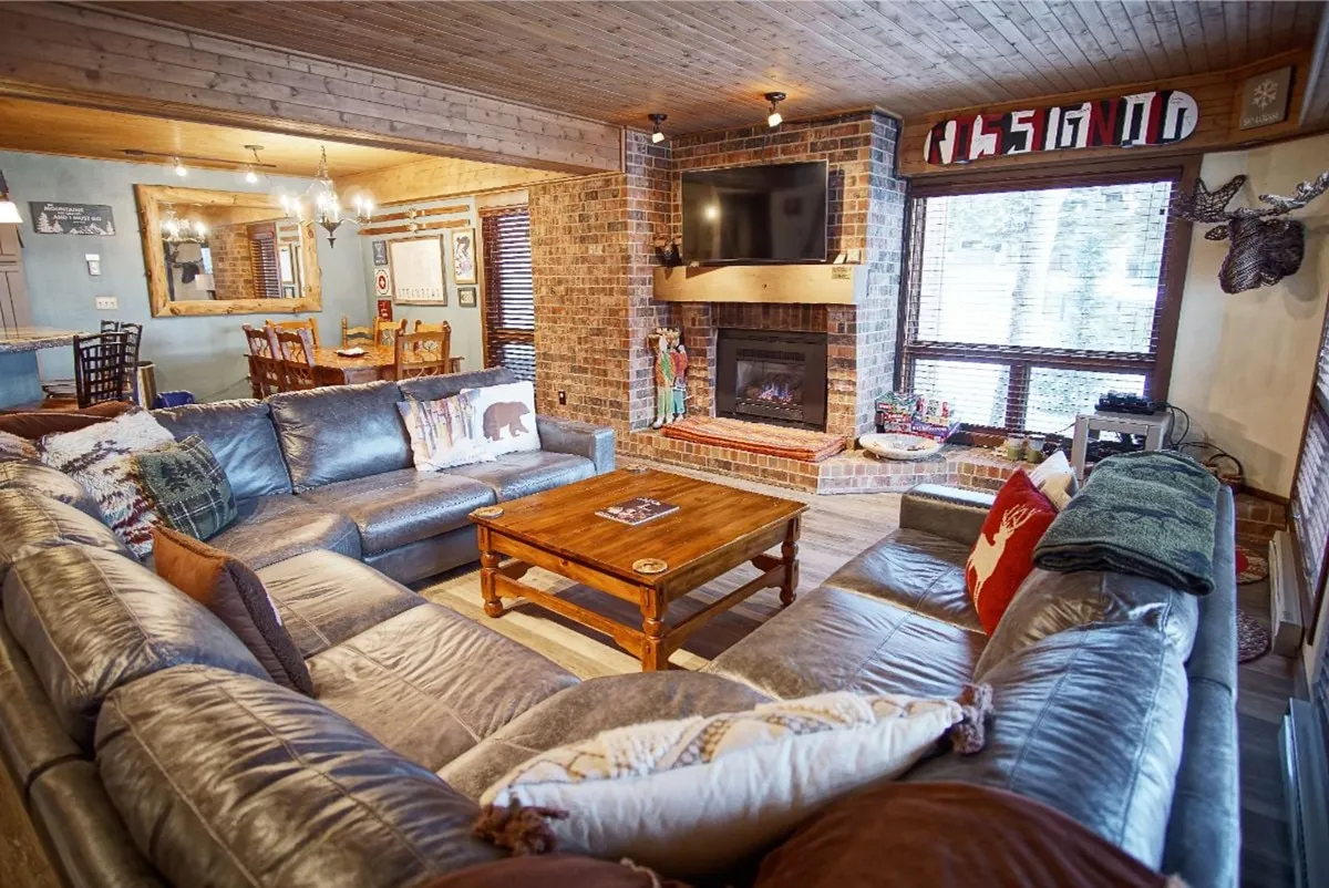 A broader view of the living area, this image captures a large sectional sofa, a glowing fireplace, a wooden coffee table, and a dining area in the background. The overall space feels open and fluid, with a clear sightline between the living and dining spaces, reinforcing the open-concept layout described in the section.
