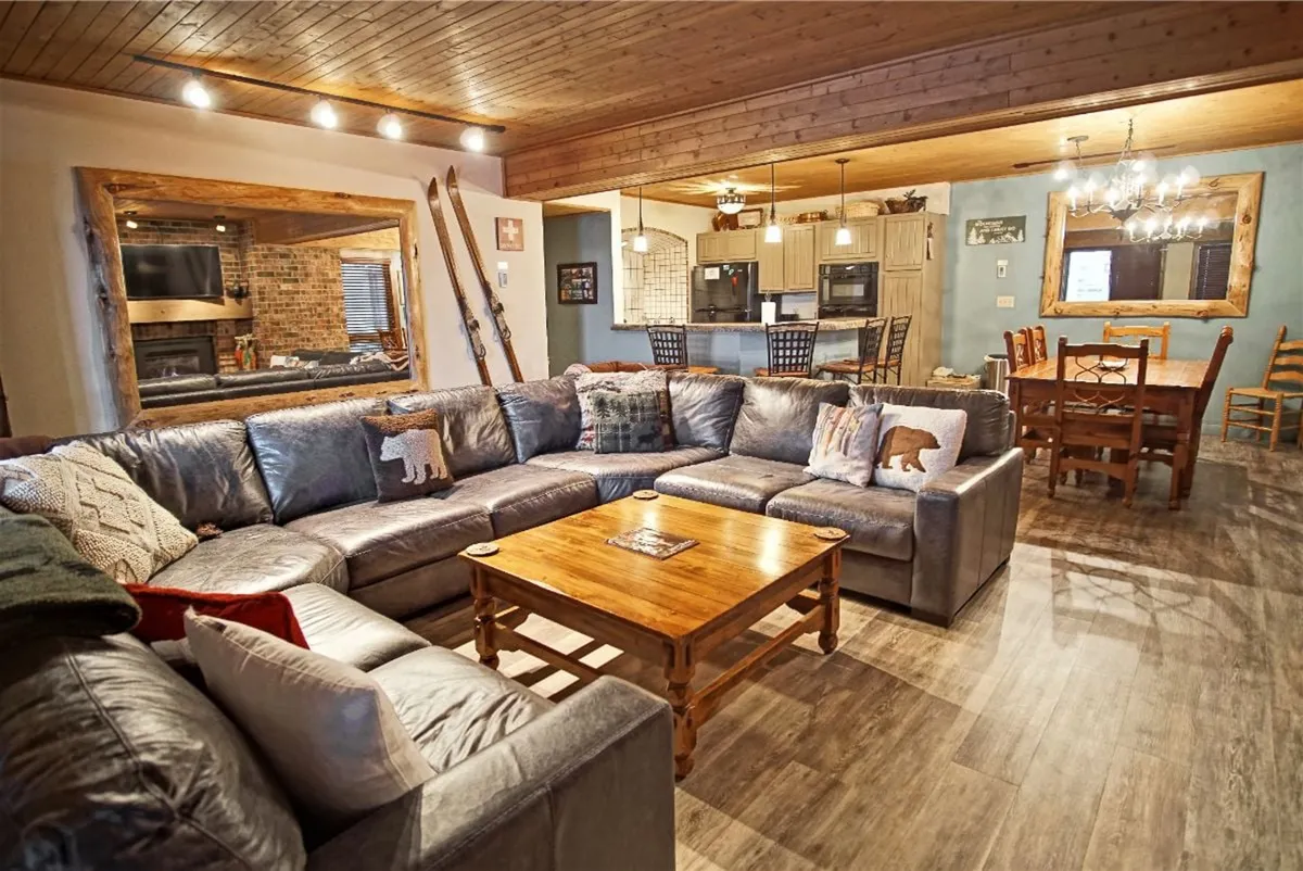 A spacious interior shot showcasing a large sectional sofa, wooden coffee table, open dining area, and a glimpse into the kitchen. The setting is warm and inviting, ideal for family gatherings, casual meals, and relaxing after a day outdoors.