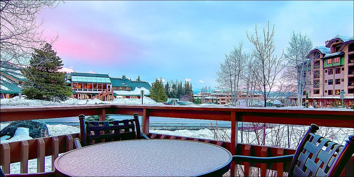An outdoor patio scene with a sturdy table surrounded by trees and distant buildings, all set atop a snow-covered ground. The space is ready for al fresco dining or relaxed gatherings in the fresh mountain air, with easy access from indoors.