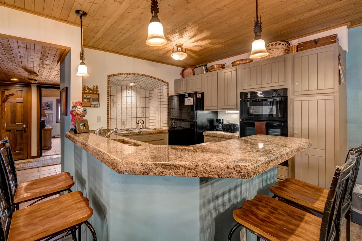 A warm and welcoming kitchen view featuring granite countertops, bar stools for casual dining or socializing, modern oven and microwave, and a decorative wall arch. The open layout encourages group participation and connection between the kitchen and adjacent spaces.