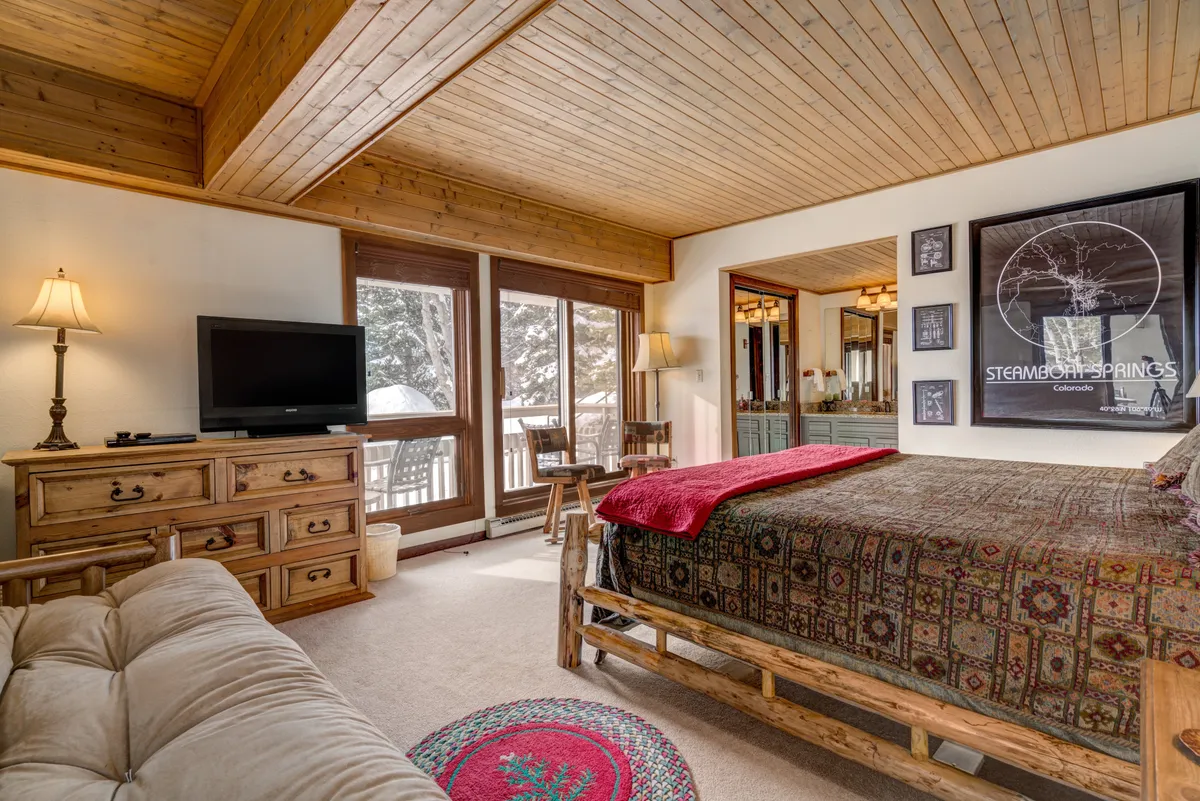 A serene bedroom with a comfortable bed featuring a patterned comforter, large windows bringing in natural light, and cozy wooden furniture. The peaceful setting invites relaxation and rest after a day exploring Steamboat.