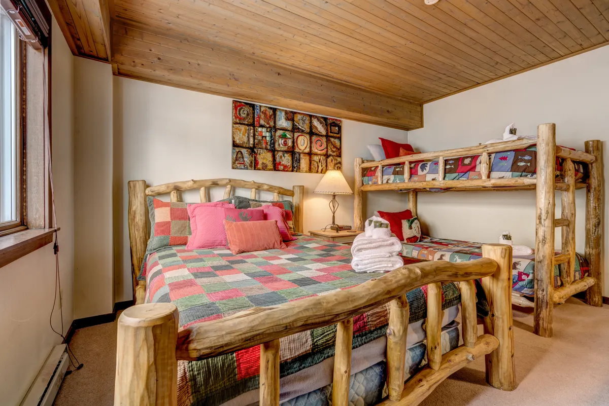 A cozy bedroom with charming log beds dressed in colorful quilts, a bedside lamp, and playful artwork. The space is perfect for families or children, offering multiple sleeping options in a warm and welcoming environment.