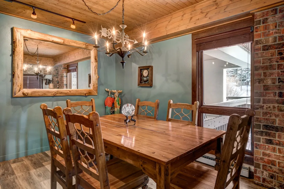 A warm, welcoming dining area with a wooden dining table set beneath a chandelier, a large mirror reflecting the space, and a brick accent wall. The setup is conducive to shared meals or memorable celebrations.