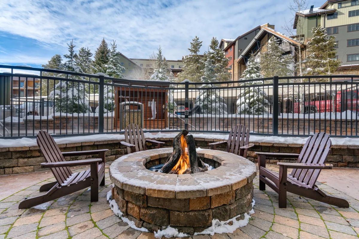 An exterior scene with a cozy fire pit surrounded by Adirondack chairs, snow-laden trees, and glimpses of surrounding buildings. The setting encourages social gatherings and warm returns after a day on the slopes.