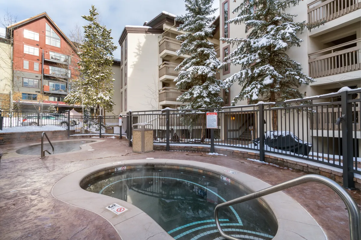 A tranquil exterior scene featuring a hot tub surrounded by snow-covered trees, with the building facade visible in the background. The wintry landscape is inviting, hinting at a welcoming retreat after a day on the slopes.