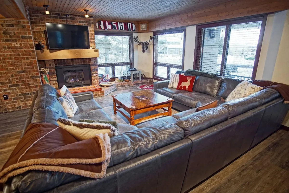 This image shows a spacious living room featuring a large sectional sofa arranged around a wooden coffee table, a prominent stone fireplace, and expansive windows that bring in natural light and highlight the mountain setting. The décor is warm and inviting, with sophisticated touches and plush furnishings creating a cozy ambiance.